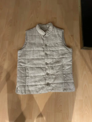 Beige quiltad John Henric linne väst med knappar - Säljer en stilren beige quiltad linneväst från John Henric med ståkrage och knäppning framtill. Västen har två sidofickor och en bröstficka, perfekt för att addera lager till din outfit. Tillverkad i ett mjukt material som känns skönt mot kroppen.