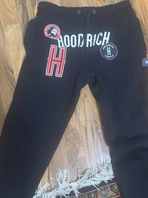 Mjukisbyxor hoodrich  - Säljer ett par svarta mjukisbyxor från Hoodrich i storlek L. Byxorna har coola patches och tryck framtill, bland annat ett stort rött H och texten HOODRICH. Dragsko i midjan för skön passform och mjukt material som är perfekt för chill dagar.