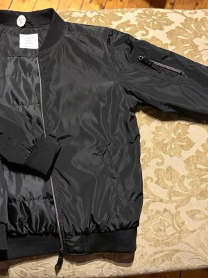 Svart bomberjacka från Lindex - Säljer en svart bomberjacka från Lindex med klassisk ribbad krage, muddar och nederkant. Jackan har dragkedja framtill, fickor på sidorna och en extra ficka med dragkedja på ena ärmen. Materialet är glansigt och känns lätt och smidigt.