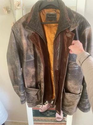 Brun skinnjacka Woodlands XL - Riktigt cool brun skinnjacka från Woodlands i storlek XL. Jackan har slitna detaljer, stora fickor och en schysst krage med fuskpäls. Insidan är fodrad i orange och har quiltad känsla. Perfekt för dig som gillar vintagevibbar och robust stil.det saknas knappar men dragkedjan funkar 