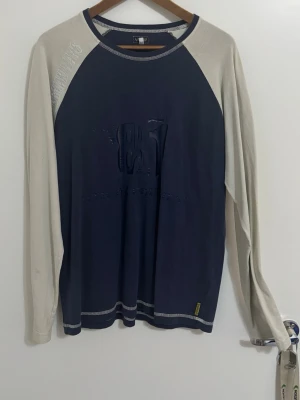 Blå och beige långärmad tröja AJ Jeans - Säljer en stilren långärmad tröja från AJ Jeans i marinblått med beige ärmar. Tröjan har tryckt 81 på bröstet och kontrastsömmar vid hals och nederkant. Perfekt för dig som gillar enkel och sportig stil.