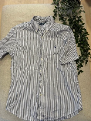Blå/vit randig Ralph Lauren skjorta M - Säljer en klassisk vit och blå randig skjorta från Ralph Lauren i custom fit.   Skjortan har kort ärm, button-down krage och den ikoniska broderade loggan på bröstet.   Tillverkad i mjuk bomull och stängs med knappar framtill.   Skjortan är i mycket bra skick förutom att den behöver en strykning 😊.   Tveka inte att höra av dig ifall du har några frågor eller liknande!