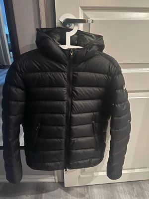 Svart Colmar pufferjacka med huva - Säljer en svart pufferjacka från Colmar med huva och dragkedja framtill. Jackan har två fickor med dragkedja och Colmar-logga på ärmen. Jackan är liten i storleken så passar S-M. 