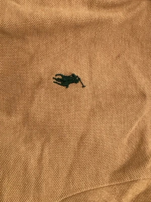 Beige pikétröja från Polo Ralph Lauren - Klassisk beige pikétröja från Polo Ralph Lauren i 100% bomull. Tröjan har korta ärmar, krage med knappar och den ikoniska gröna broderade loggan på bröstet. Perfekt för en chill och stilren look.