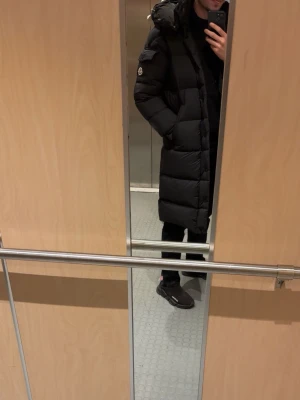 Svart lång dunjacka från Moncler - Säljer en svart lång dunjacka från Moncler med huva och stora quiltade partier. Jackan har en klassisk Moncler-logga på ärmen och praktiska fickor. Perfekt för kalla dagar och riktigt snygg till streetwear-stil.