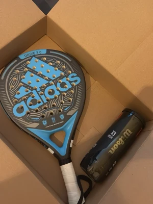 Ingen sko synlig på bilderna - Det finns inga skor på bilderna, endast en Adidas padelracket och Wilson padelbollar i en kartong. Ingen information om skor kan ges.
