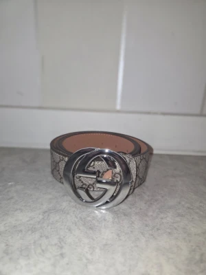 Gucci GG Supreme bälte med silverdetalj - Säljer ett klassiskt Gucci GG Supreme bälte med ikoniskt silverfärgat GG-spänne. Bältet är beige med brunt monogrammönster och har en lyxig känsla. Perfekt accessoar för att lyfta vilken outfit som helst.