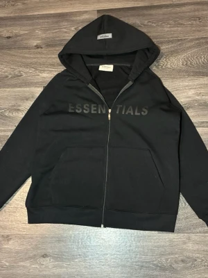 Essentials Zip - Säljer en stilren svart hoodie från Essentials med dragkedja. Klassisk och trendig design som passar perfekt till både vardag och streetwear. Använd fåtal gånger och väl omhändertagen. Fraktar direkt efter köp. Pris kan diskuteras vid snabb affär. 