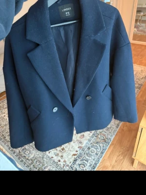 Mörkblå kavajjacka från Lindex XS - Säljer en mörkblå kavajjacka från Lindex i återvunnen ullmix. Jackan har dubbelknäppning med två knappar, breda slag och två fickor framtill. Passformen är loose och modellen är lite boxig, perfekt till lager på lager under höst och vinter.