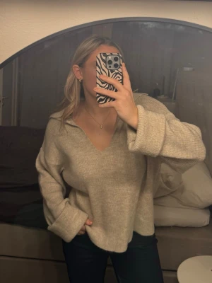 Beige stickad tröja från Pieces - Supermysig beige stickad tröja från Pieces med v-ringning och bred krage. Tröjan har långa ärmar med uppvik och lite oversized. Perfekt att slänga på sig under hösten eller vintern.