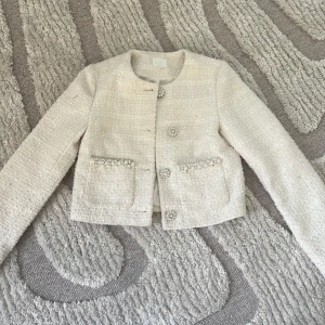 Vit pärlbroderad kofta i bouclé - Superfin beige vit i bouclé med pärl- och strassbroderade knappar och fickor. Croppad modell med lång ärm och rund halsringning. Perfekt att styla med jeans för en chic look. Materialet är strukturerat och ger en lyxig känsla.💕