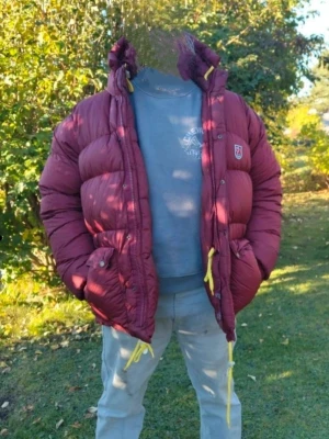 Vinröd dunjacka från Fjällräven M - Säljer en vinröd, fluffig dunjacka från Fjällräven i storlek herr M. Jackan har huva, gula dragskor och dragkedja framtill. Perfekt för kalla dagar och riktigt skön att bära. Klassisk Fjällräven-logga på bröstet och riktigt snygg passform. Den har slitage på dragskon på blixtlås. Jag har storlek XL annars på jackor så denna är stor M. Fruktansvärt varm. Perfekt nu!  Behöver rensa garderob så köp gärna 😀👍🏼
