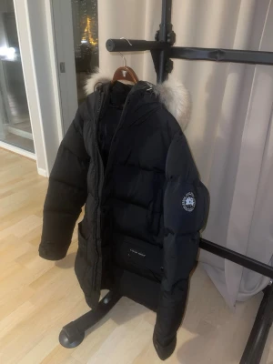 Svart Canada Goose dunjacka med päls - Säljer en svart dunjacka från Canada Goose med stor fluffig päls runt huvan. Jackan har dragkedja, stora fickor och den exclusiva nya patch på ärmen. Perfekt för kalla dagar och riktigt varm. Passar dig som vill ha en stilren och funktionell vinterjacka. (Kan gå ner i pris)