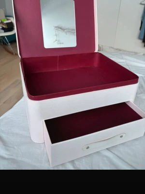 Sminklåda från CAIA - Säljer en snygg sminklåda från CAIA i ljusrosa med vinröd insida och inbyggd spegel. Väskan har ett stort fack och en utdragbar låda, perfekt för att organisera smink och borstar. 