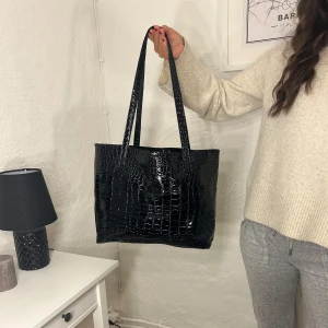 Svart väska - Snygg svart axelväska med krokodilmönster. Väskan har en praktisk dragkedja och rymligt inre med vinrött foder. Stor och får plats med allt du behöver inför dagen!