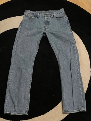 Levi's 501 ljusblå jeans W30 L30 - Klassiska Levi's 501 jeans i ljusblå tvätt. Rak passform med normal midja och fem fickor. Tillverkade i slitstarkt bomullsjeansmaterial. Perfekta för en avslappnad och tidlös look. Ikonisk läderpatch bak i midjan och knappgylf framtill.
