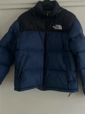 Blå och svart pufferjacka The North Face - Säljer en blå och svart pufferjacka från The North Face med broderad logga på bröstet. Jackan har hög krage, dragkedja framtill och två fickor med dragkedja. Perfekt för kalla dagar och har en klassisk streetstil. original price 2500, strl 36/38 (S) gender neutral expandable hood, well suited for rain and/or snow used well so the arms have lost slight amount of “puff” but still very warm and comfortable, looks normal:) 