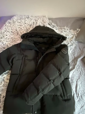 Canada goose Wyndham black label 2025 - Säljer en svart pufferjacka från Canada Goose i storlek M. Jackan har huva och avtagbar fuskpäls, dragkedja och knappar framtill samt två stora fickor. Perfekt för kalla dagar och riktigt snygg streetstil.