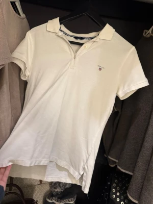 Vit pikétröja från Gant, stl S - Klassisk vit pikétröja från Gant i storlek S. Tröjan har korta ärmar, krage med två knappar och diskret Gant-logga broderad på bröstet. Tillverkad i mjuk bomull som känns skön mot huden. Perfekt till jeans eller kjol för en clean look.
