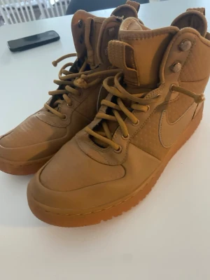 Hög NIKE skor  - Snygga nike  sneakers i helbrunt med matchande snörning och gummisula. Skorna har högt skaft, klassisk Nike-logga på sidan och grovt mönstrad yttersula för extra grepp. Små skavmärken på skaftet, se bild uppifrån.