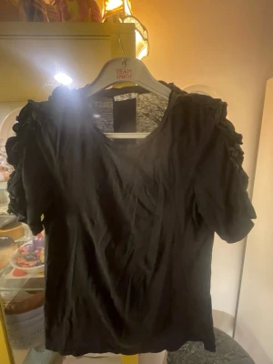 Svart t-shirt med volangärmar Stl M-L - Svart t-shirt från Zara Trafaluc med korta ärmar och snygga volangdetaljer på axlarna. T-shirten har en enkel rund halsringning och är gjord i ett mjukt material.