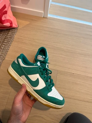 Nike Dunk Low gröna och vita sneakers - Säljer ett par Nike Dunk Low sneakers i grönt och vitt med klassisk swoosh på sidan. Ovandelen är i mocka och textil, med gröna snören och gummisula i beige. Perfekta för dig som gillar streetwear och vill ha en stilren sneaker med retrovibbar.