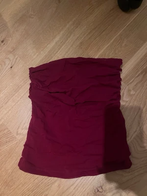 Vinröd bandeau tubtopp - Säljer en vinröd bandeau tubtopp i mjukt bomullsmaterial. Toppen är axelbandslös och har en enkel, stilren design med lätt rynkning upptill för en snygg passform. Perfekt att matcha med jeans eller kjol för en cool look.