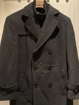 Mörkgrå skepparkavaj från Ralph lauren - Stilren mörkgrå skepparkavaj i ull från Ralph Lauren som tyvärr blivit för liten för mig. Dubbelknäppt med bred krage, innerfoder och ribbad hög krage med dragkedja. Klassisk design med fickor och snygga knappar framtill. Perfekt för kalla dagar när du vill se extra sharp ut. 