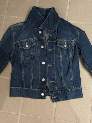 Mörkblå jeansjacka med hög krage - Säljer en mörkblå jeansjacka från Diesel med hög krage och silverfärgade knappar. Jackan har två bröstfickor med lock och knapp, samt markerade sömmar för en klassisk look. Perfekt till vår och höst för en cool och avslappnad stil.