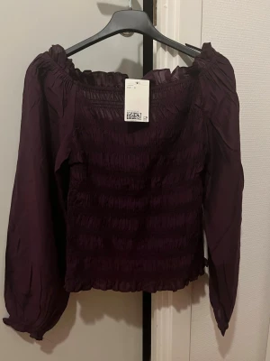 Purple Smocked Off-Shoulder Top – Size S (New with Tag) - Beautiful deep purple off-shoulder top with long sleeves and a stretchy smocked fit. The fabric is lightweight and comfortable, with a flattering silhouette that works both dressed up and casual. Never worn, still with tag attached. Perfect for everyday wear, dinner, or a night out. Size: S Condition: New with tag Color: Dark purple / plum Fit: Stretchy, fitted Feel free to message me if you have any questions 🌸 