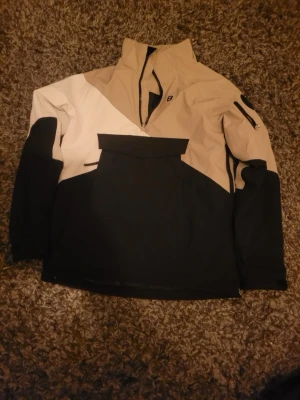 Beige och svart vindjacka med half zip - Säljer en snygg beige, svart och vit vindjacka från Bergans. Jackan har en praktisk half zip, hög krage och stor ficka framtill. Ärmarna har dragkedjeficka och jackan är perfekt för blåsiga dagar. Materialet känns slitstarkt och sportigt.