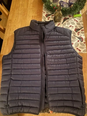 Save The Duck väst - Säljer en marinblå dunväst med horisontella quiltade sömmar och hög krage. Västen har en smidig dragkedja framtill och är perfekt att slänga över en hoodie eller tröja för extra värme. Enkel och stilren design som funkar till det mesta.