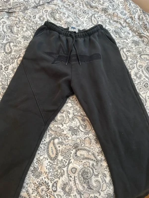 Svarta mjukisbyxor från NOFS - Svarta mjukisbyxor från NOFS med broderad logga framtill. Byxorna har resår och snörning i midjan för skön passform och är gjorda i mjukt bomullsmaterial. Perfekta för chill dagar eller när du vill vara bekväm men ändå se snygg ut.