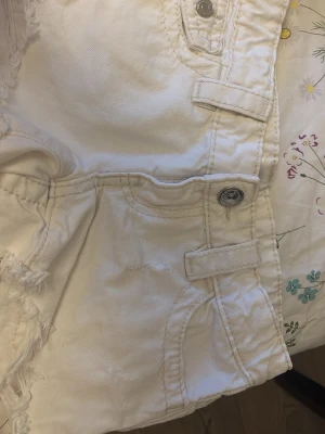 Vita fransiga jeansshorts - Säljer ett par vita jeansshorts med fransiga kanter och coola detaljer som fickor med lock och silvriga knappar. Shortsen har bälteshällor och är perfekta för varma dagar. De är tillverkade i ett mjukt bomullstyg som känns skönt mot huden.