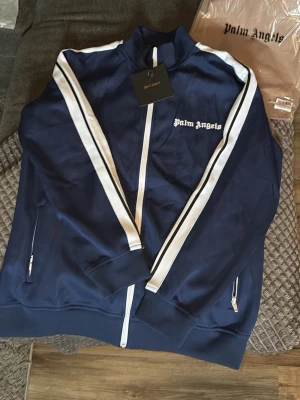 Blå hoodie från Palm Angels - Snygg blå hoodie från Palm Angels med vita och svarta ränder längs ärmarna. Jackan har dragkedja framtill, ribbade muddar och Palm Angels-logga broderad på bröstet. Perfekt för dig som gillar streetwear och vill sticka ut.