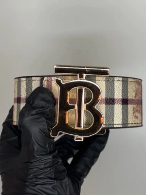 Rutig Burberry bälte med B-spänne - Säljer ett klassiskt Burberry bälte med det ikoniska rutiga mönstret i beige, svart och vinrött. Bältet har ett stort, guldigt B-spänne som verkligen sticker ut. Perfekt accessoar soppar upp vilken outfit som helst.