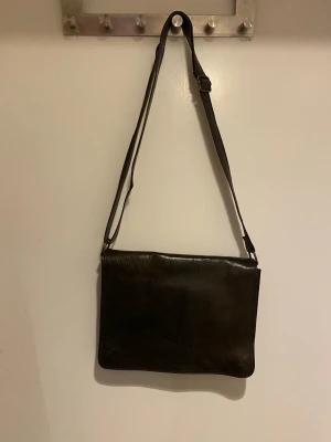 Mörkbrun axelväska i skinn - Snygg mörkbrun axelväska i äkta skinn med justerbar rem och magnetknäppning. Väskan har flera fack, inklusive ett med dragkedja, vilket gör den praktisk för att hålla ordning på dina grejer. Perfekt storlek för att bära med sig det viktigaste och passar till många olika stilar.