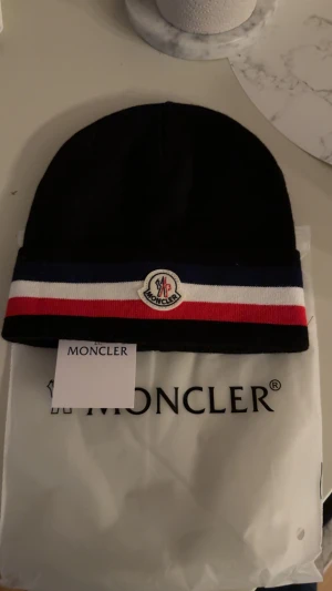 Moncler mössa  - Snygg svart mössa från Moncler med klassiska ränder i blått, vitt och rött runt kanten. Mössan har Moncler-logga framtill och är tillverkad i mjukt material som sitter skönt på huvudet. Perfekt för dig som vill ha en stilren accessoar med lyxig känsla.