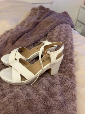 Vita sandaler med klack från SANTIA - Säljer ett par stilrena vita sandaler från SANTIA med breda korslagda remmar och öppen tå. Skorna har en stadig klack och justerbart spänne vid ankeln. Perfekt för dig som gillar enkel och fräsch design med lite höjd.