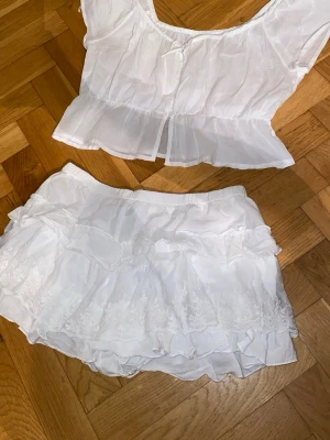 Vit blus och skort set  - Supersöt vit blus med tillhörande skort från Nelly. Blusen har korta ärmar, rund halsringning med knyt och en luftig, flowy passform. Perfekt för sommarens varma dagar och ger en romantisk vibe tillsammans med matchande skort.