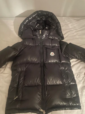 Svart Moncler pufferjacka med huva - Svart pufferjacka från Moncler med glansig finish, huva och dragkedja framtill. Jackan har Moncler-logga på bröstet, praktiska fickor med dragkedja och ett coolt serietidningsmotiv på insidan. Perfekt för kalla dagar och riktigt snygg streetstil.