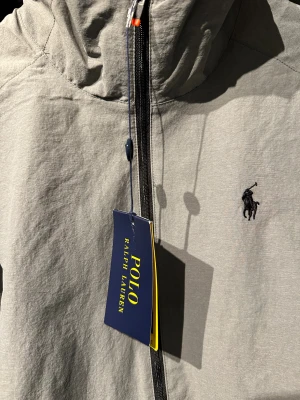 *helt ny* vind/skidjacka från Ralph Lauren - Snygg grå vindjacka från Polo Ralph Lauren med klassisk logga på bröstet. hög krage för halsskydd. Helt nya oanvänd. köpt för 1700 kr
