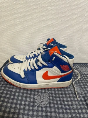 Nike Air Jordan 1 blå och vit - Nike Air Jordan 1 sneakers i blått och vitt med orange detaljer och logga. Skorna har snörning, rund tå och platt sula. Ovandelen är i skinn och har perforeringar på tån för extra ventilation. Snygg kontrast mellan färgerna och klassisk Jordan-vibe.