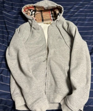 Grå Burberry hoodie S. MEN L KVINNOSTORLEK - Snygg grå hoodie från Burberry i storlek S.Men L i kvinnostorlek! Tröjan har dragkedja framtill och en huva med klassiskt Burberry-rutigt foder i beige, svart och rött. Mjukt material och ribbade muddar vid ärmar och nederkant. Perfekt för dig som gillar stilrena märkesplagg.