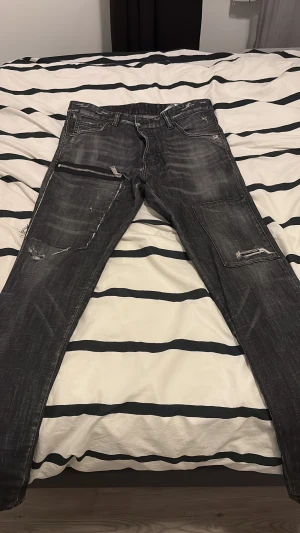 Svarta Dsquared2 jeans med dragkedjor - Säljer ett par svarta jeans från Dsquared2 i storlek 48. De har slitna detaljer, dragkedjor på benen och en cool, tvättad look. Byxorna har raka ben och klassiska fickor både fram och bak. Materialet är jeans med inslag av polyester för extra komfort.