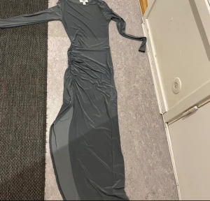 Grå långklänning Rebecca Stella XS - Snygg grå långklänning från Rebecca Stella i storlek XS. Klänningen har lång ärm, tight passform och är gjord i ett mjukt syntetmaterial. Den har en cool rynkad detalj längs sidan och en slits nedtill som ger extra edge.