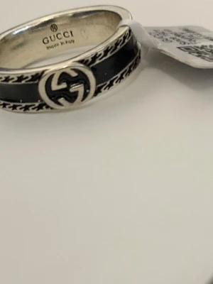 Gucci silverring med svart emalj - Säljer en snygg ring från Gucci i  silver med svart emalj och det klassiska GG-logot i mitten. Ringen har detaljerade mönster längs kanterna. Gick den i present men har aldrig använt den då storleken är fel.