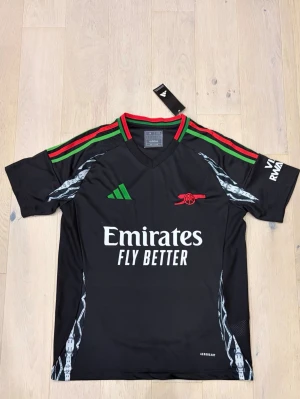 Arsenal x Adidas svart fotbollströja M - Svart Arsenal fotbollströja från Adidas med röda och gröna ränder på axlarna, broderat rött kanonmärke och vit Emirates Fly Better-tryck. Slim fit och tillverkad i polyester med mönstrade detaljer på sidorna. Perfekt för dig som älskar fotboll och snygg design.