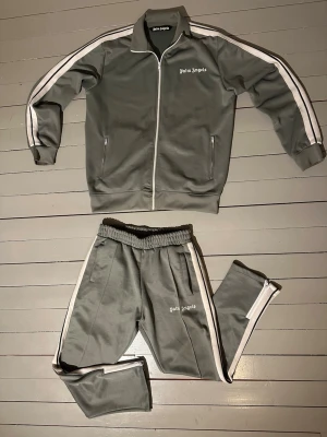 Palm Angels grå tracksuit - Helt ny, oanvänd Palm Angels tracksuit i grått med vita ränder längs ärmar och ben. Setet består av zip-jacka med ståkrage och matchande byxor med elastisk midja och dragkedja vid benslut. Palm Angels-logga tryckt på bröstet och benet. Snygg och sportig look med klassiska detaljer.