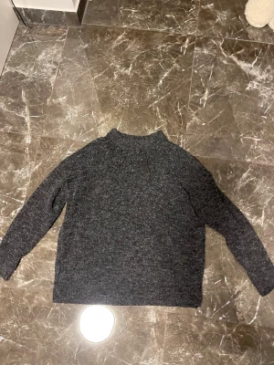 Mörkgrå stickad polotröja 157 - Mysig mörkgrå stickad polotröja från 157 i storlek M. Tröjan har en hög krage och långa ärmar, perfekt för kyliga dagar. Materialet är grovstickat och ger en skön och avslappnad känsla. Enkel att matcha med jeans eller mjukisbyxor.